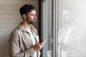 A man is standing in front of a window, looking at his cell phone. He appears focused on the screen, with natural light coming through the window. photo
