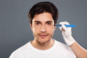 Cosmetic Operation. Portrait Of young man before plastic surgery, doctor hands with scalpel near patient's face photo