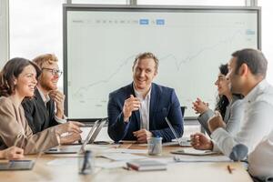A group of business professionals are gathered around a conference table in a modern office. The group is engaged in a conversation and appears to be working together on a project photo