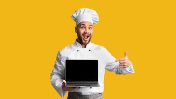 Excited Chef Cook Man Showing Laptop Empty Screen And Gesturing Thumbs-Up Approving Culinary Website Standing On Black Studio Background. photo