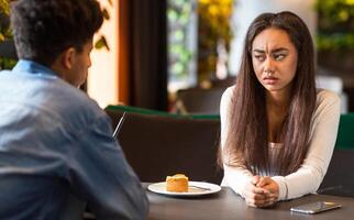 African American young girl with long hair and a white shirt sits at a table, looking concerned, while a guy in a blue shirt talks to her in a modern cafe. photo
