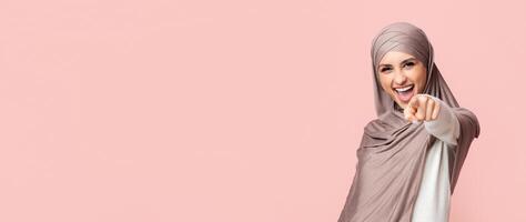 Ha ha ha. Muslim girl in hijab laughing and pointing finger at camera, mocking on somebody over pink background with empty space photo