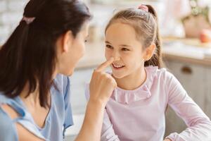 A mother and her young daughter are playing a game where they touch each others noses. The girl is smiling and looking at her mother with happiness. They appear to be in their kitchen photo