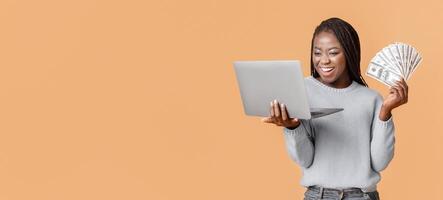 A joyful young African American woman is smiling broadly as she holds a stack of cash in one hand and a laptop in the other. She appears to be celebrating a financial achievement, copy space photo