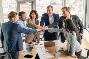 A diverse group of business professionals stand around a conference table, their hands stacked together in a gesture of teamwork and unity. The office is modern and bright photo