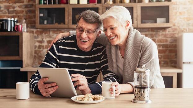un mayor Pareja es sentado juntos a un mesa en su cocina. el hombre es mirando a un tableta mientras el mujer es sonriente y mirando a a él. ellos Aparecer a ser compartiendo un momento de alegría y la risa foto