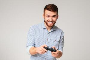 Excited millennial guy playing games with joystick and smiling, isolated on grey studio background, free space photo