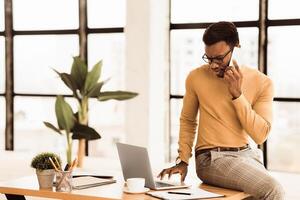 Solving Issue. Confused afro businessman talking on phone at workplace, using laptop, sitting on desk. Copy space photo