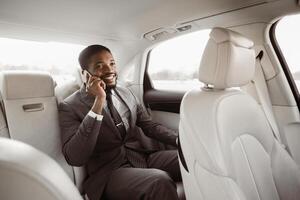 Black businessman in a suit is sitting in the back seat of a car while talking on his phone. He has a serious expression on his face and is looking to the side photo