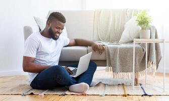 A young black man sits on the floor of his living room, using a laptop computer. He is sitting on a rug in front of a couch, with a plant on a side table photo