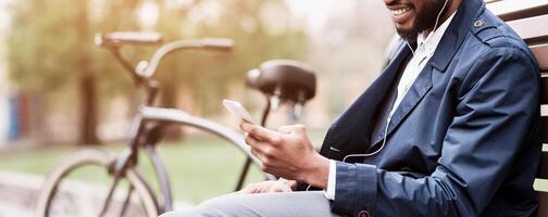 A businessman is seated on a park bench, using his smartphone, with a bicycle nearby. The environment is serene and professional, showcasing a balance between work and nature, cropped photo