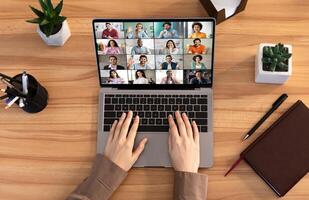 A person is participating in a conference using a laptop computer. The screen shows multiple people in a grid format. The persons hands are on the keyboard of the laptop, top view photo