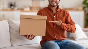 Cropped of man is sitting on a white couch in a living room, holding a cardboard box in front of him with his left hand. He is smiling and gesturing with his right hand photo