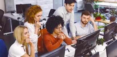 A group of focused multiethnic colleagues are seated at desks using desktop computers in a well-lit, contemporary office. They appear to be engrossed in work, displaying concentration and teamwork photo