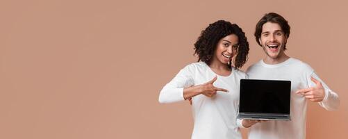 Cool website. Smiling multiracial couple holding laptop with black screen and pointing at it, posing together over yellow background with empty space photo