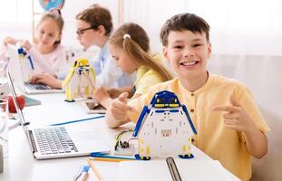 A young boy sits in a classroom, beaming with pride as he points to a robot he built. He is surrounded by other students who are also engaged in STEM activities. photo