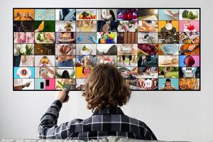 A man sits on a couch, facing a television screen, watching a montage of images. The screen is filled with a grid of various pictures, each depicting a different scene or activity. photo