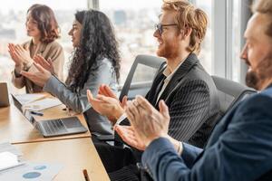 A group of business professionals sit at a table in a modern office, clapping their hands in appreciation as they listen to a presentation. They are dressed in formal attire photo