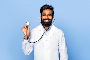 A smiling Indian doctor with a beard holds up a stethoscope against a light blue background. He is wearing a white lab coat and appears confident. He is likely working in the medical field photo