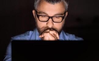 A man with glasses is intently concentrating while working on his laptop in a dimly lit room. His hands are clasped together under his chin as he focuses on the screen. photo