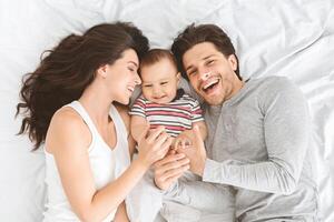 A young couple is lying on a white bed, smiling at their baby. The father is on the right side of the image and the mother is on the left. The baby is lying in the middle of the bed photo