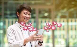 Asian young man stands in an urban setting, smiling as he scrolls through his smartphone. He is surrounded by glowing pink notification bubbles with white hearts inside, indicating likes photo