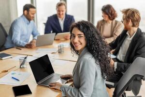 A young woman with curly dark hair smiles at the camera while sitting at a conference table with three other people. She is working on a laptop and the others are using tablets and computers. photo