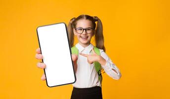 Smartphone And School. Excited Elementary Student Girl Pointing Finger At Empty Phone Screen Over Yellow Background. Empty Space, Mockup photo