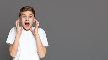 An elated teenager with hands near face and wide eyes, isolated on a grey background, showing shocked expression, copy space photo