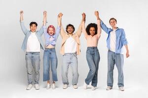 A group of five diverse young adults stand in a studio setting with their arms raised, holding hands and smiling joyfully. They are dressed casually in jeans and shirts. photo