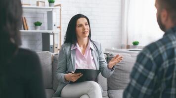A lady therapist sits on a gray couch in a modern office and talks to a man client. She gestures with her right hand while holding a clipboard photo