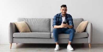 Cool Gadget And Application. Portrait of young smiling Arab man holding mobile phone, typing sms message, sitting on the couch in living room. Guy browsing internet, surfing web, using app, free space photo