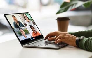 Cropped of person is participating in a conference on a laptop. They are sitting at a table and using their hands to type on the keyboard. The laptop screen shows four people in a chat. photo
