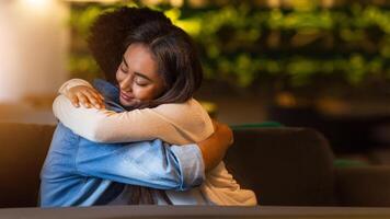 Two black friends share a heartfelt embrace inside a cozy cafe with soft, dim lighting. The background features lush, green decor which adds to the warm atmosphere of the evening setting. photo