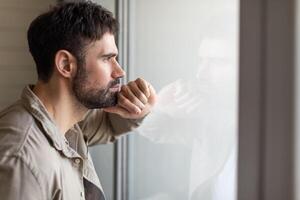 A man stands near a window, gazing outside with his hand resting on his face. He appears contemplative and deep in thought as he observes the view before him. photo