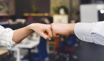 A close-up shows two individuals engaging in a fist bump, a gesture of affirmation or greeting, with one wearing a striped shirt. The background suggests an informal office environment, cropped photo