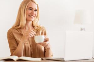 A woman sits at a desk with a laptop and an open notebook, smiling as she enjoys a cup of coffee. photo
