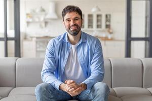 A man with a beard, wearing a blue shirt and jeans, sits on a gray couch in a modern living room. He smiles warmly at the camera, with his hands clasped in his lap. photo