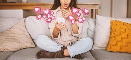 A young woman, sitting on a couch, is looking at her smartphone. She is surrounded by animated, pink heart-shaped notifications. The woman appears to be scrolling through social media feed, cropped photo
