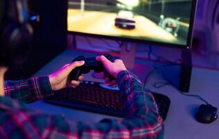 This image shows a gamer playing a racing game at their computer desk. The gamer is wearing a plaid shirt and using a black game controller. The computer monitor is displaying racing game photo