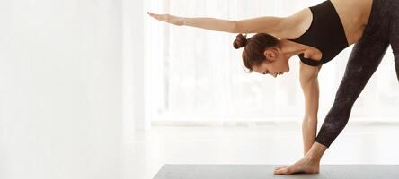 A woman is practicing a yoga pose on a mat indoors. She is focused and balancing on one leg with arms extended. The room is well-lit, and she is surrounded by a calm and minimalistic setting. photo