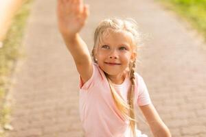 A young girl with blonde hair and blue eyes is shown reaching out with her right hand. She is wearing a light pink t-shirt and is looking directly at the camera with a warm smile, closeup photo