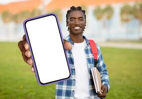 A cheerful young black man with braided hair carrying a red backpack, holds out a smartphone with a blank screen while standing on a university campus with buildings in the background photo