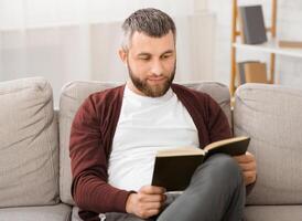 Bearded man is seated comfortably on a couch, engrossed in a book he is reading. The room is quiet as he flips through the pages, his focus solely on the words in front of him. photo