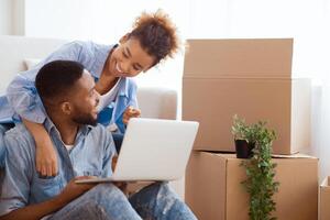 Excited Afro Spouses Using Laptop Computer Sitting On Sofa Among Unpacked Moving Boxes In New House. Free Space, Selective Focus photo