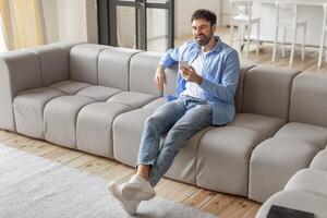 A man, dressed in jeans and a light blue shirt, sits on a light grey sofa in a modern living room. He is smiling as he uses his smartphone, copy space photo