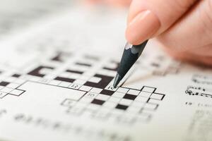 A person is seen actively engaged in a crossword puzzle, holding a pencil and carefully filling in the boxes with letters. The crossword grid is visible, showing a mix of empty and filled squares. photo
