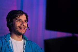 A young man smiles while looking off to the side. He is wearing a blue shirt and a headset. He is sitting in front of a computer with a purple and blue backdrop. photo