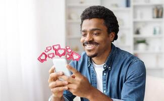 Black man with a joyful expression smiles as he looks at his smartphone. He is clearly pleased with the notifications he is receiving, which are represented by heart-shaped icons floating above photo