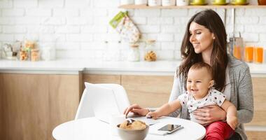 A woman is sitting at a kitchen table, holding a baby in her lap. She is working on a laptop computer, with a smartphone and a bowl of snacks in front of her, copy space photo